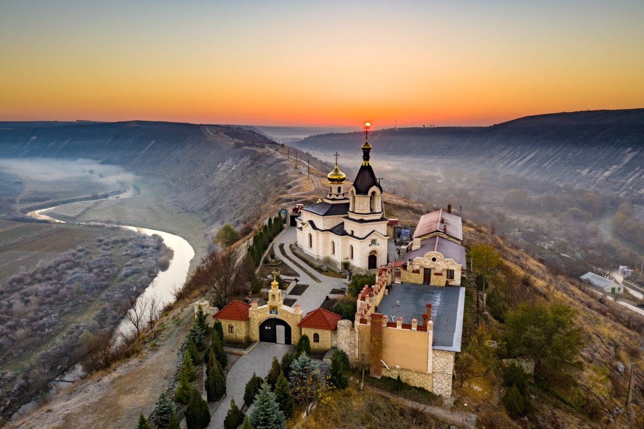 Orheiul Vechi Monastery on a mountain with courtyard and several buildings, limestone cliffs and river in the background, fog, sunrise 