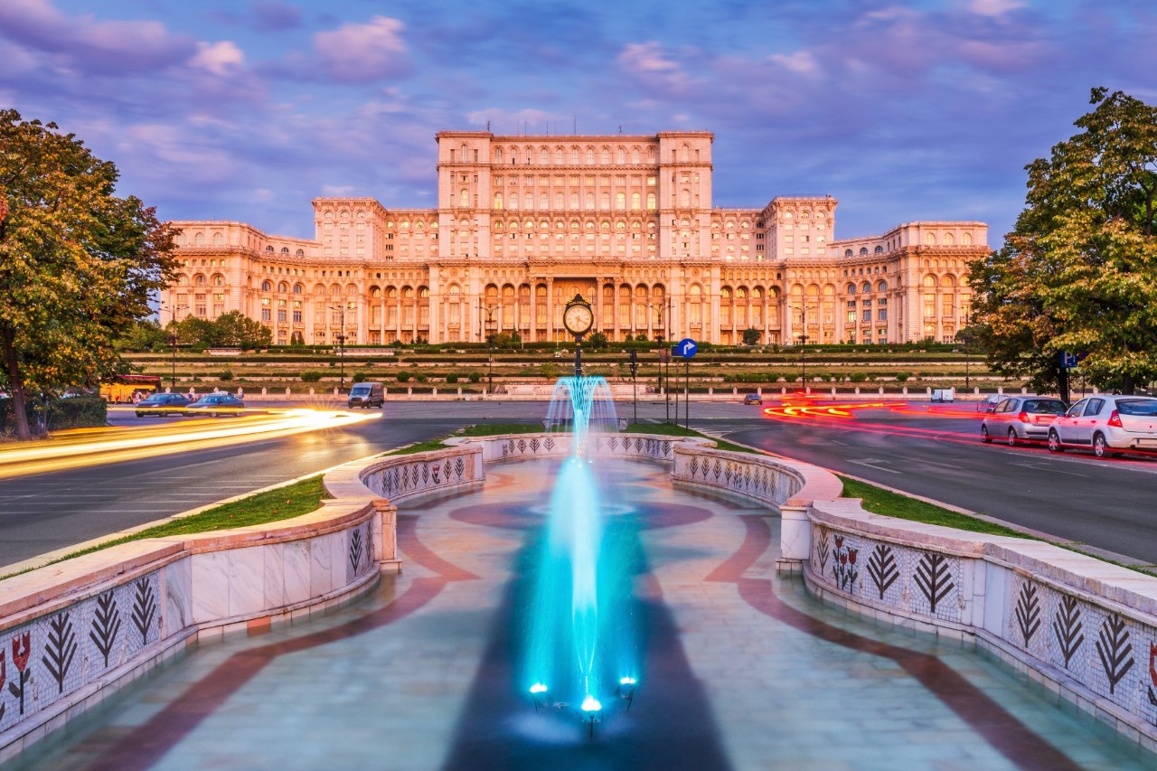 View of the monumental parliament building with illuminated fountain in the foreground, street, cars and trees. 