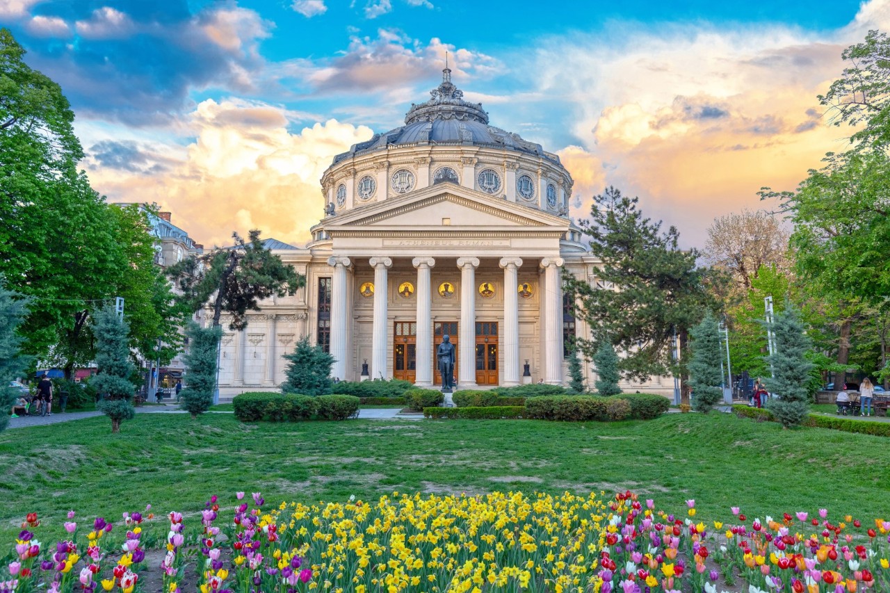 Wide park with flower beds, lawns and hedges leading to a large white building with a dome and six Greek columns. There is a statue in front of the building and tall trees at the sides. 
