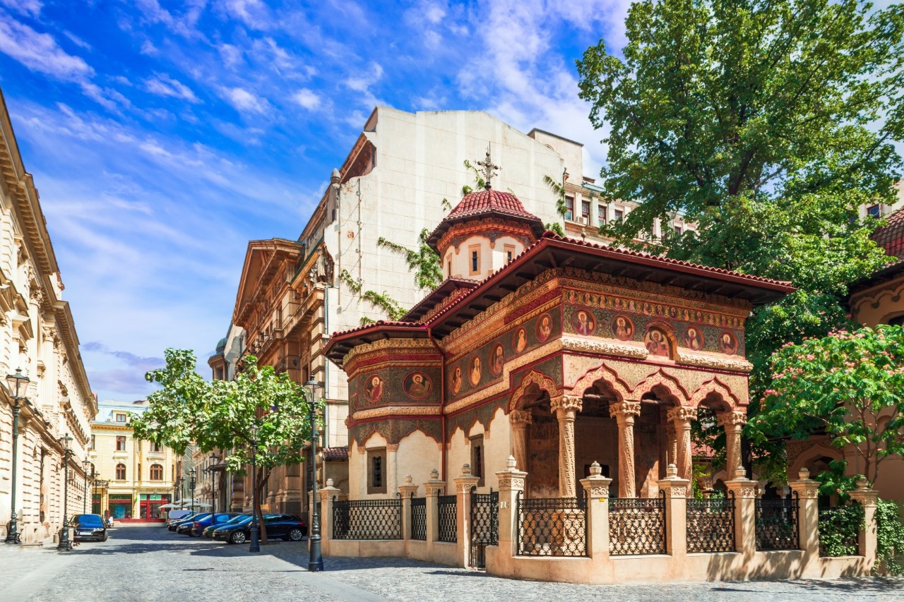 Deserted street with parked cars, buildings, trees and old, red-coloured church with dome, embellishments and arches in the foreground. 