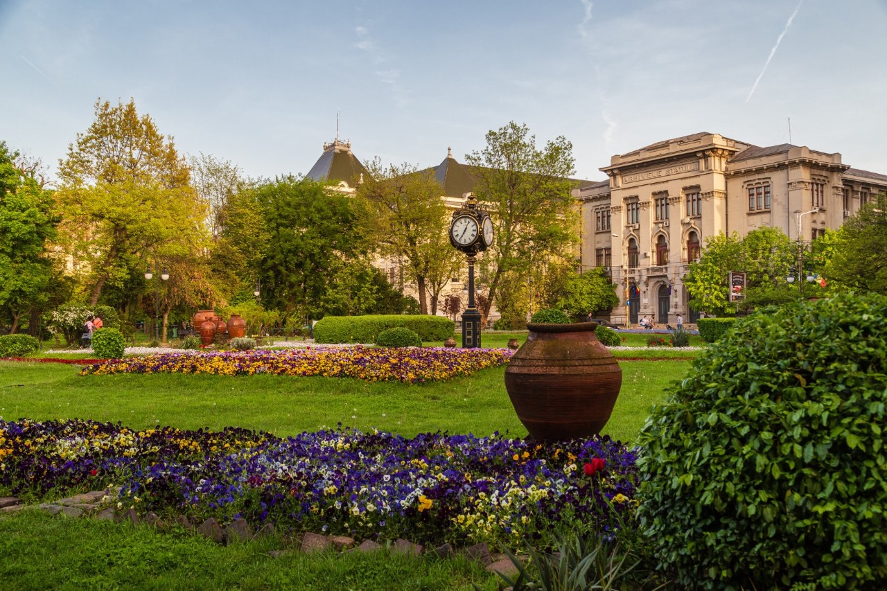 Park with flower beds, bushes, trees, grandfather clock and a building in the background. 