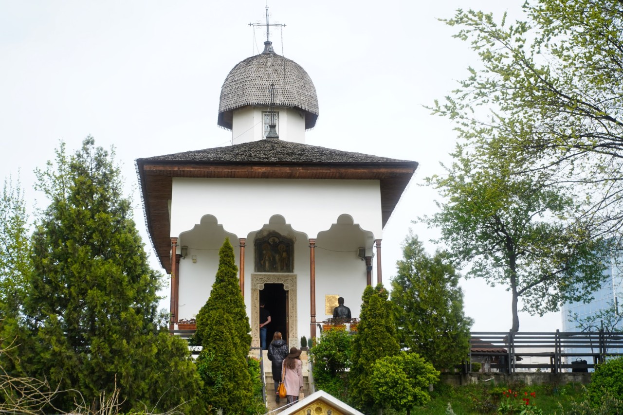 White, narrow church with some people at the entrance and trees in front of it. 