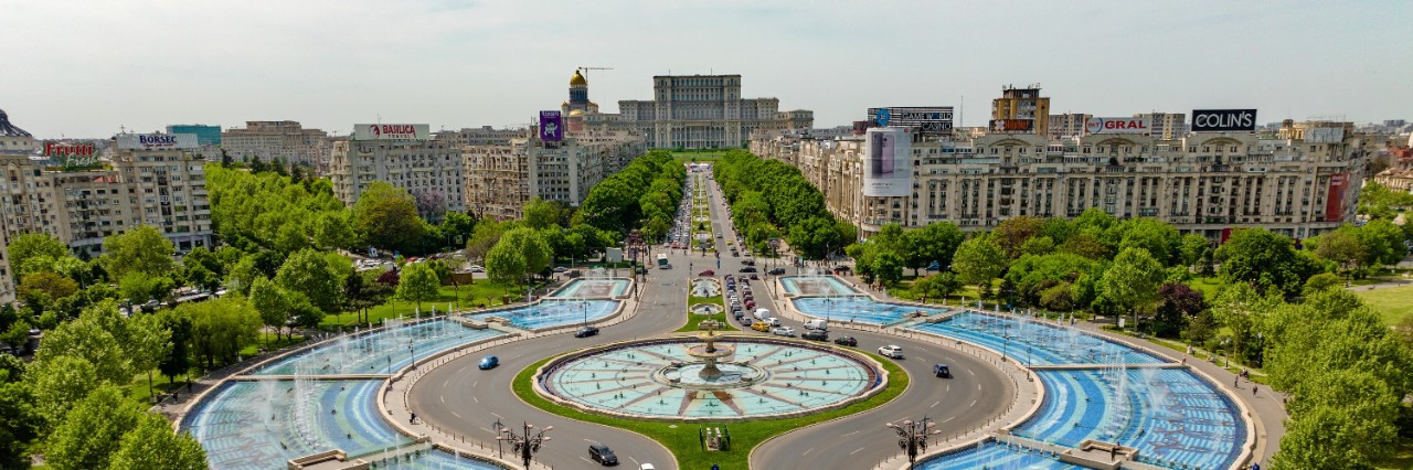View from above of Bucharest’s Unirii Square with a wide boulevard, fountains, cars, trees, buildings and the Palace of the Parliament in the background. © xpabli/stock.adobe.com    