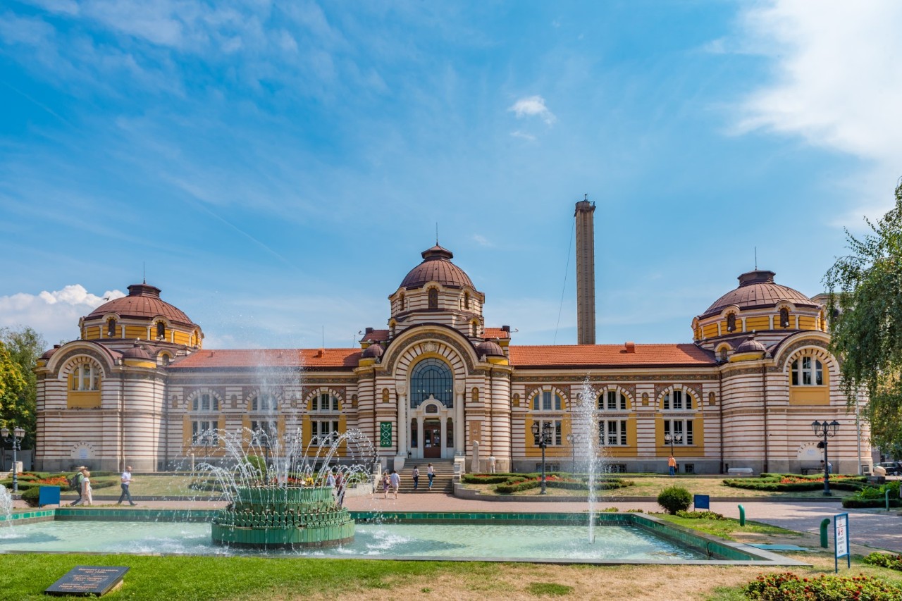 The picture shows a magnificent, elongated, historic building with a colourful façade and red roofs. It has three domes. A chimney rises in the background. In front of the building is a large rectangular fountain and some people milling around.