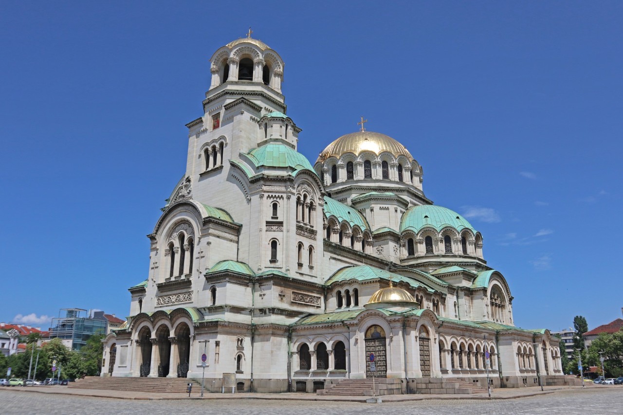The picture shows an imposing cathedral with light green and golden domes against a blue sky. The façade of light-coloured stone is decorated with round arches, columns and reliefs. There is a square around the cathedral where people can be seen.