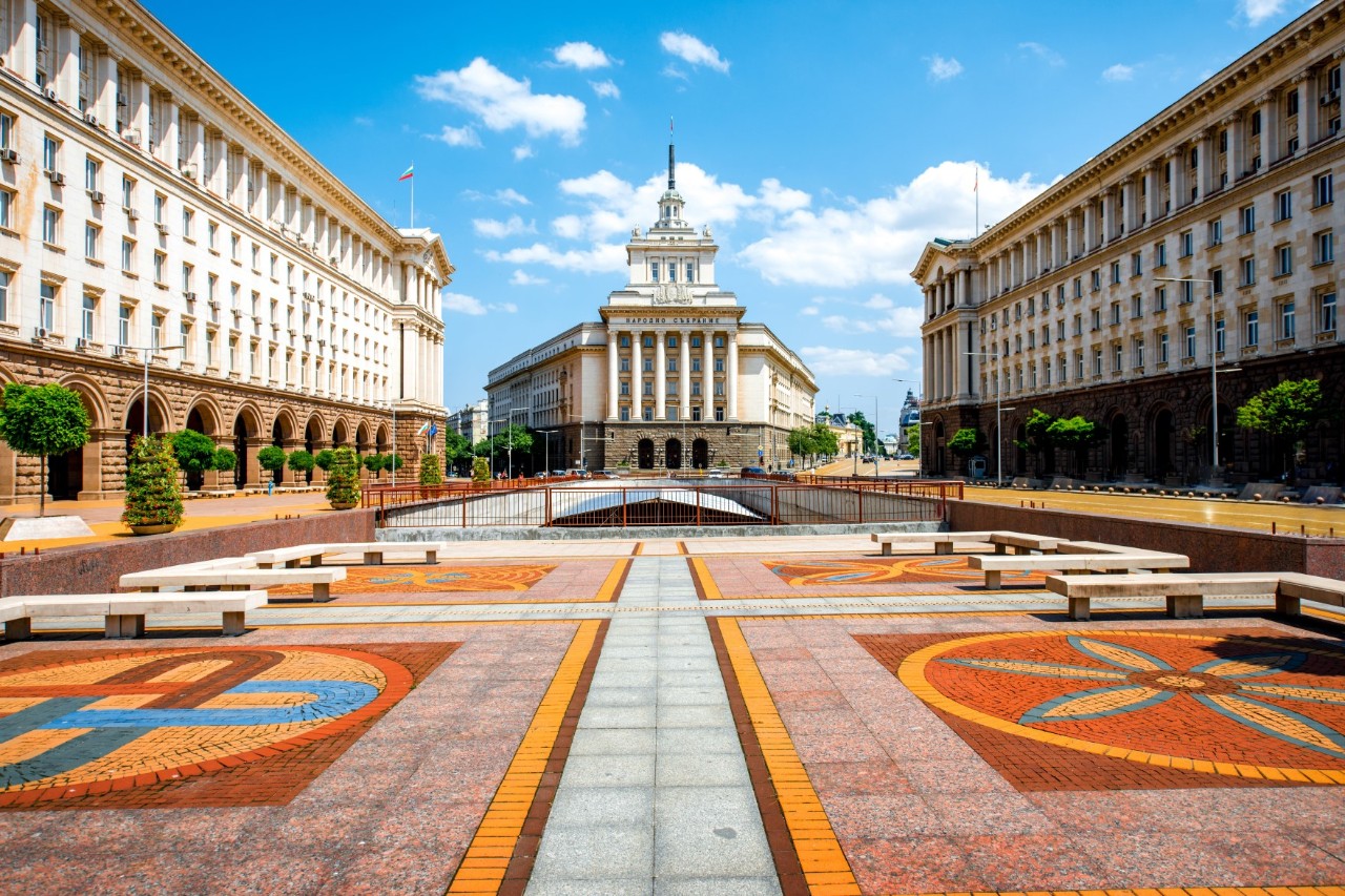 The image shows a large central square paved with large rectangular stone slabs in a regular, geometric pattern. The patterns are coloured, and the dominant shade is orange. Symmetrically on either side of the square is a large building built in the socialist classicist style. In the centre is a third building of the same design with a central tower. The sky is blue with white clouds.