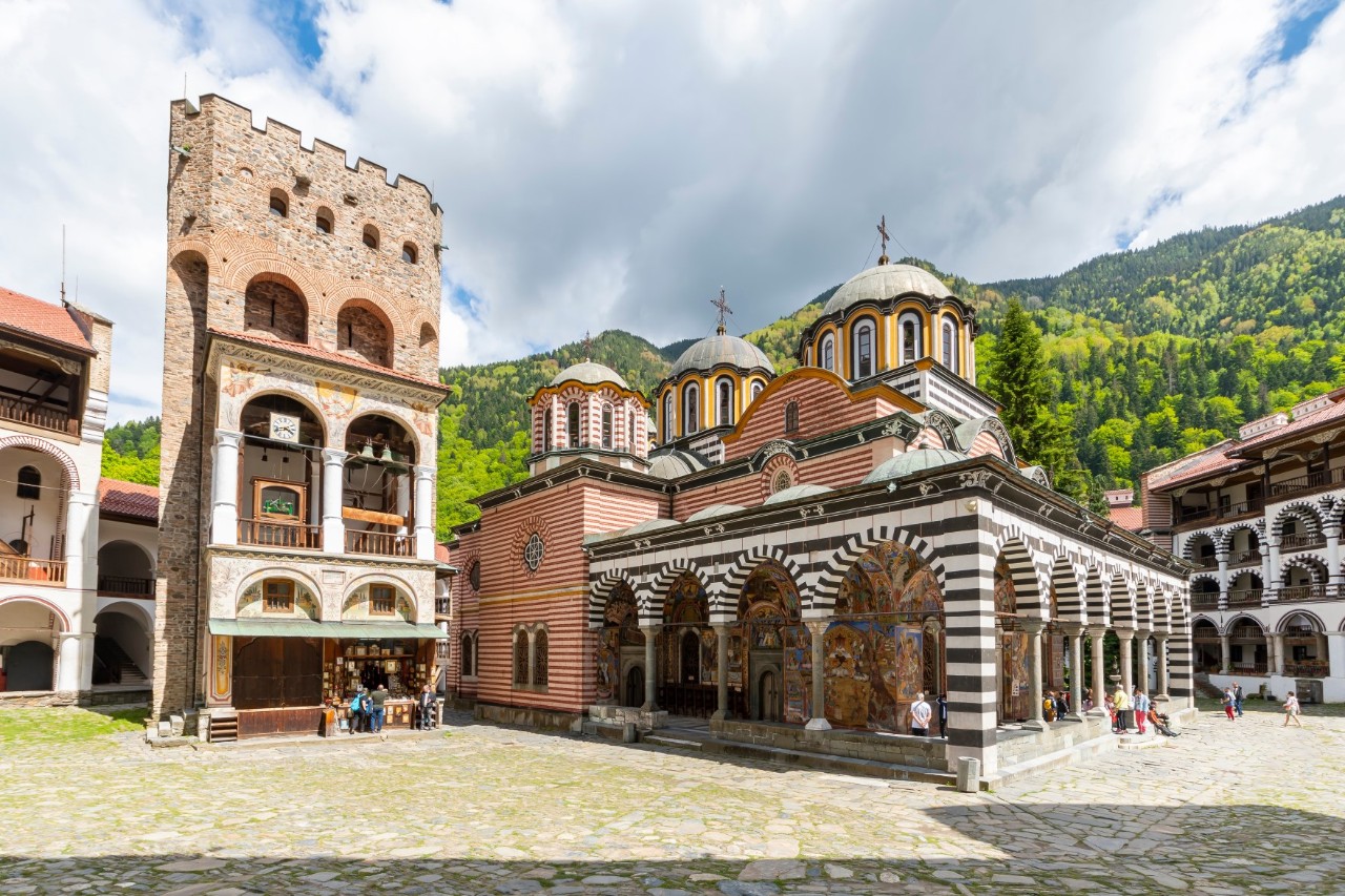 The picture shows the inner courtyard of a monastery. In the foreground, on a large square, is the main church with striking black and white stripes on the arches and porticoes as well as several domes. To the right of the church are richly decorated arcades. On the left of the picture is a high square defence tower. A green, wooded mountain landscape rises in the background. The sky is cloudy. A few people can be seen scattered around the courtyard.