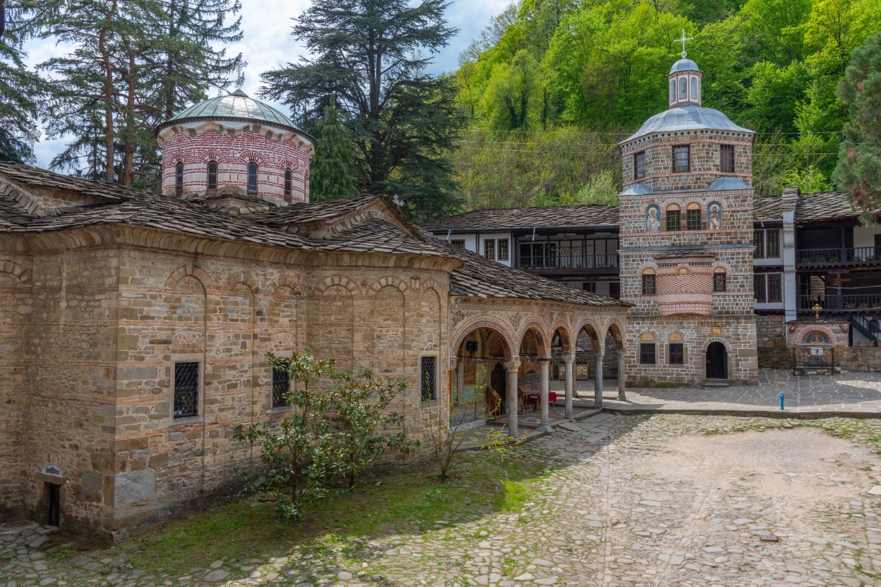 Pictured is a historic monastery set in green surroundings. The buildings are made of natural stone and have slightly arched roofs, domes and ornate entrances with round arches. A cobbled square can be seen in front of them.