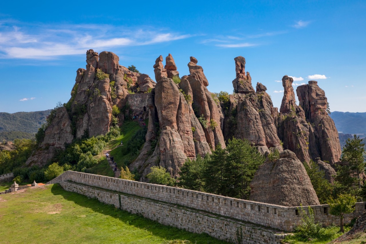 View of a rock formation with towering brown rocks against a blue sky. In front of the group of rocks is a fortress wall and a green meadow. Several green trees grow between the rocks. The sky is blue.