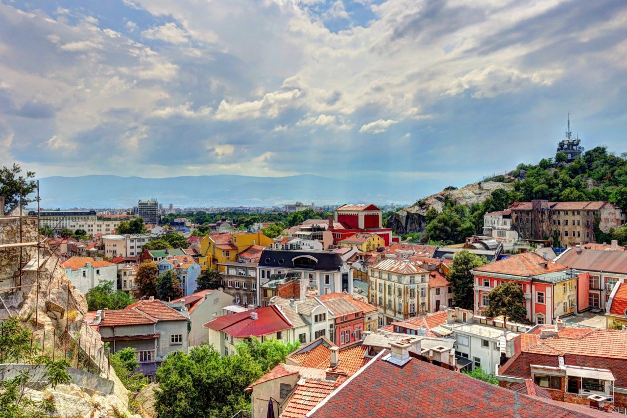 View over the roofs of a city with colourful houses on a sunny day. In the background on the right-hand side of the picture is a green hill on which a kind of tower with an antenna can be seen. A rocky outcrop can be seen on the left. In the far distance in the background is a mountain range that can only be vaguely recognised. The sky is bluish-purple cloudy.