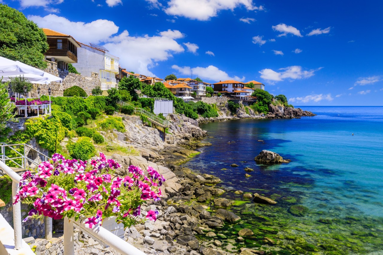 The picture shows a seaside resort with turquoise-coloured water. In the foreground, blooming pink flowers adorn a railing, while rocky cliffs and green terraces run behind it. Houses with red roofs stand on the cliffs.
