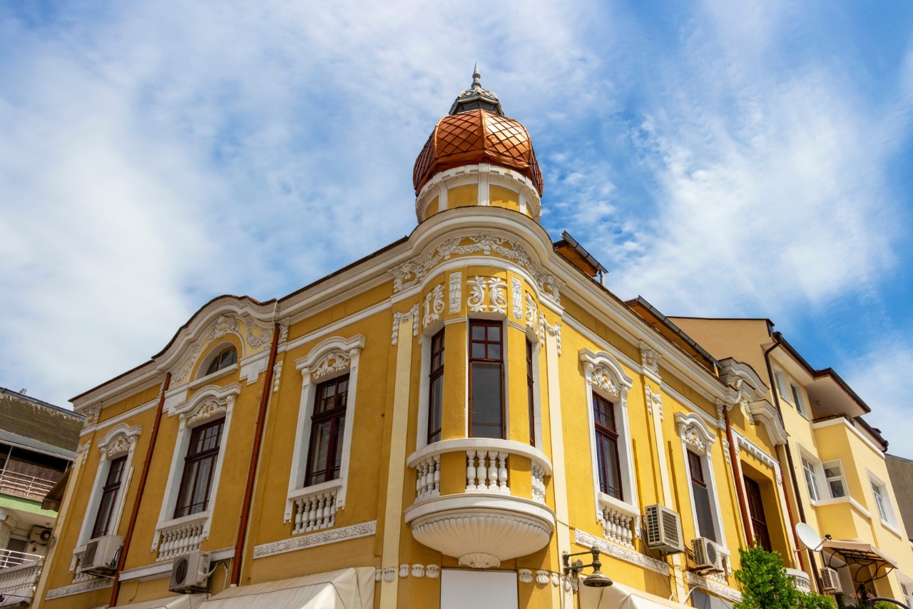 The picture shows a historic yellow corner building with decorative white stucco elements and a striking round tower with a copper-coloured dome. The sky is blue and slightly cloudy.