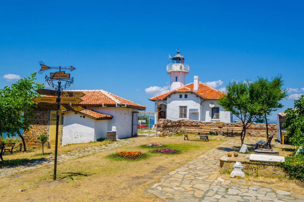 The picture shows a square with a white lighthouse and buildings with red tiled roofs. A path leads through a well-kept environment with trees, flowers, benches and a signpost. 