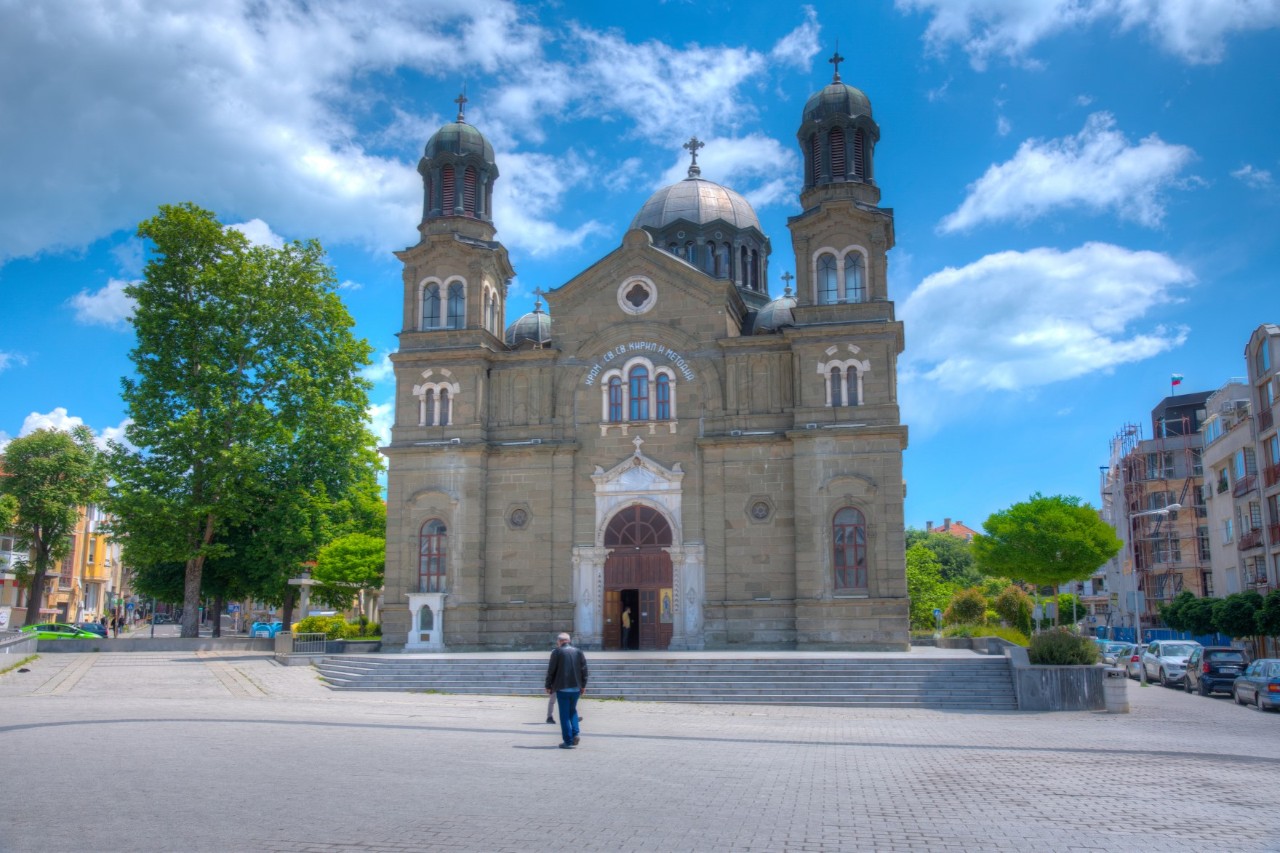 The picture shows a church with two towers and a central dome. It stands on a paved square, surrounded by green trees and houses. A person approaches the entrance to the church.