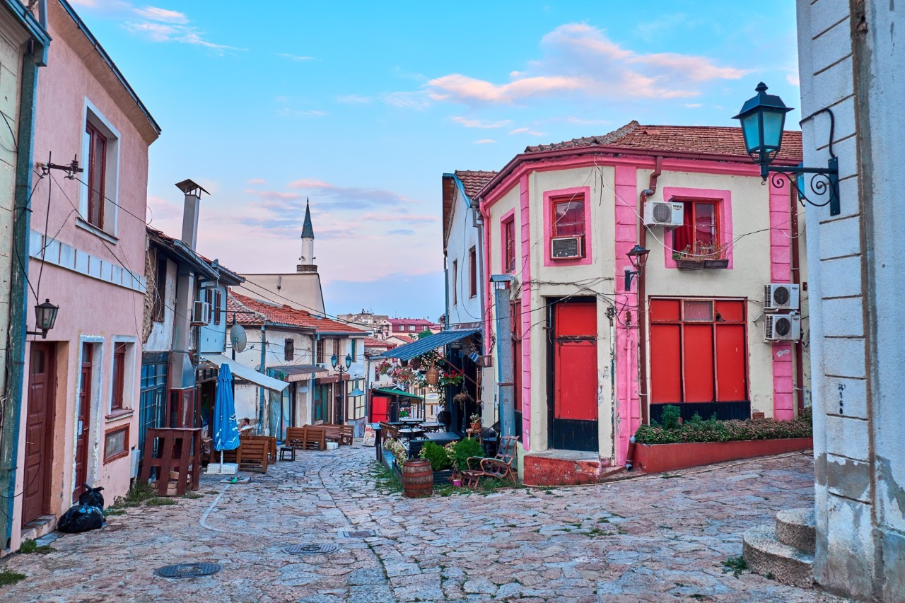 Deserted cobblestone street with predominantly small, pink buildings. A pointed tower be seen in the background.