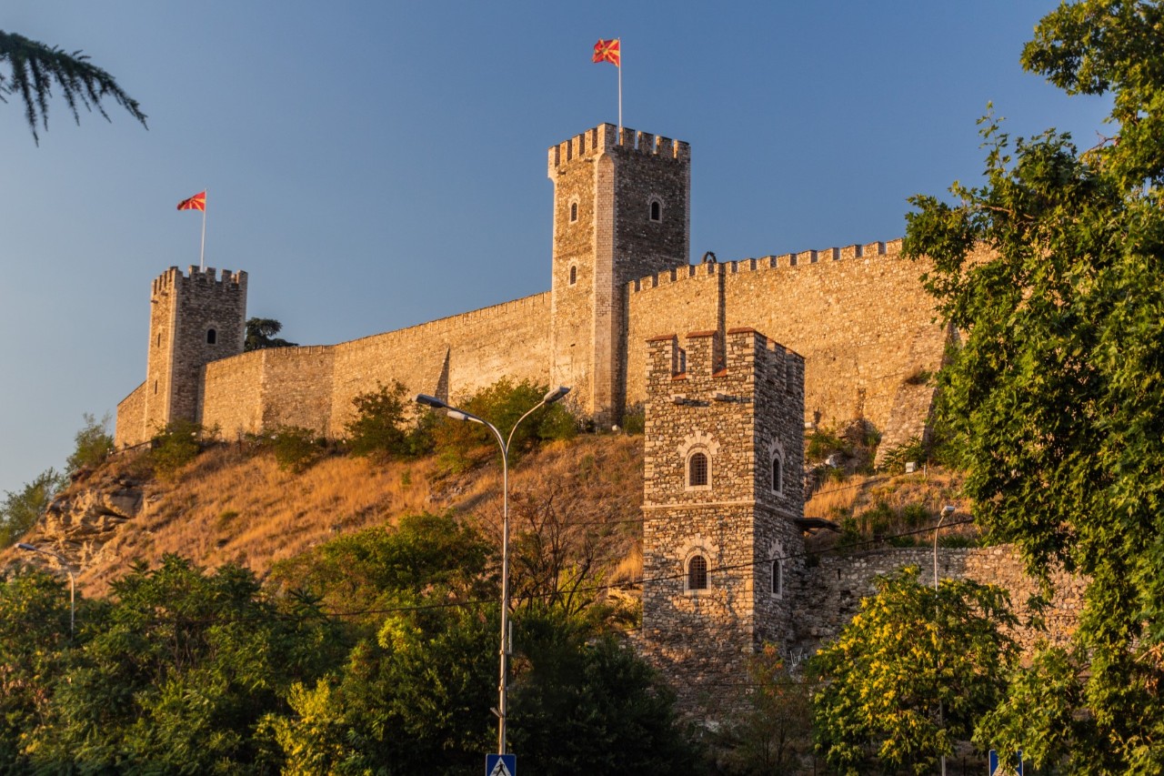View from below of a fortress on the hill with a wall and three watchtowers, each flying a flag. The fortress is surrounded by green shrubs and trees.