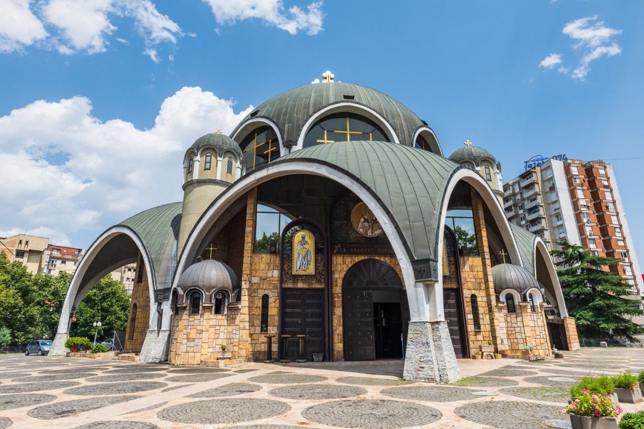 View of a striking church building that resembles a mosque with domed roofs. Golden crosses can be seen in the windows of the uppermost dome. There are multi-storey residential buildings behind the church.