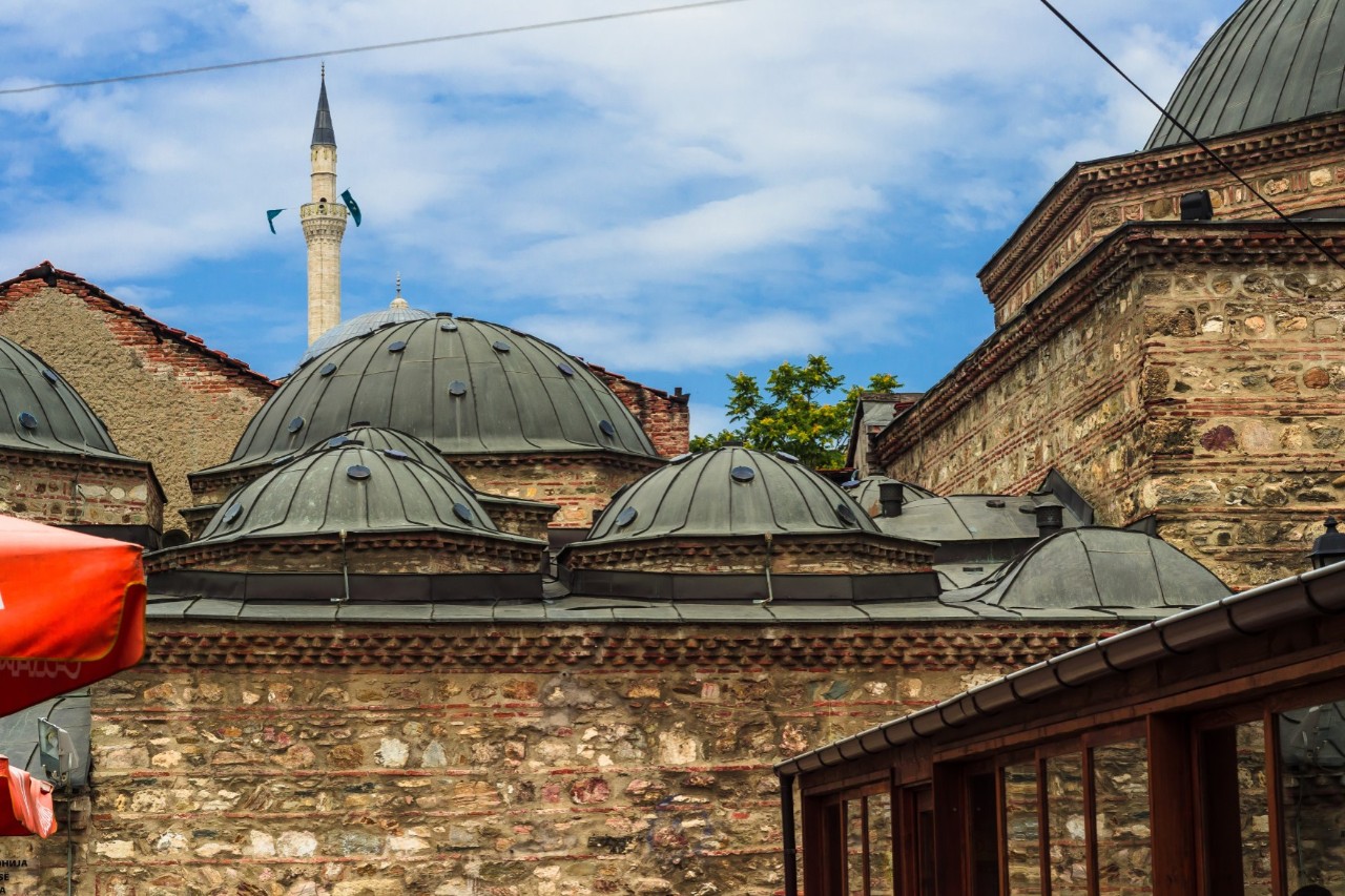 View of the upper part of an old stone building with several small green domes on the roof. A pointed tower (minaret) can be seen in the background.