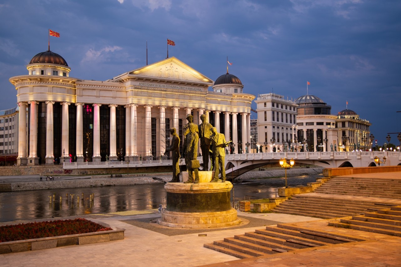 View of a white, striking building with many columns, which stands directly on the river, over which a light-coloured bridge runs. Flags are flying on the roof. There are other white buildings to the right of the museum building. In the foreground is a riverside promenade with steps and a platform with sculptures of people.