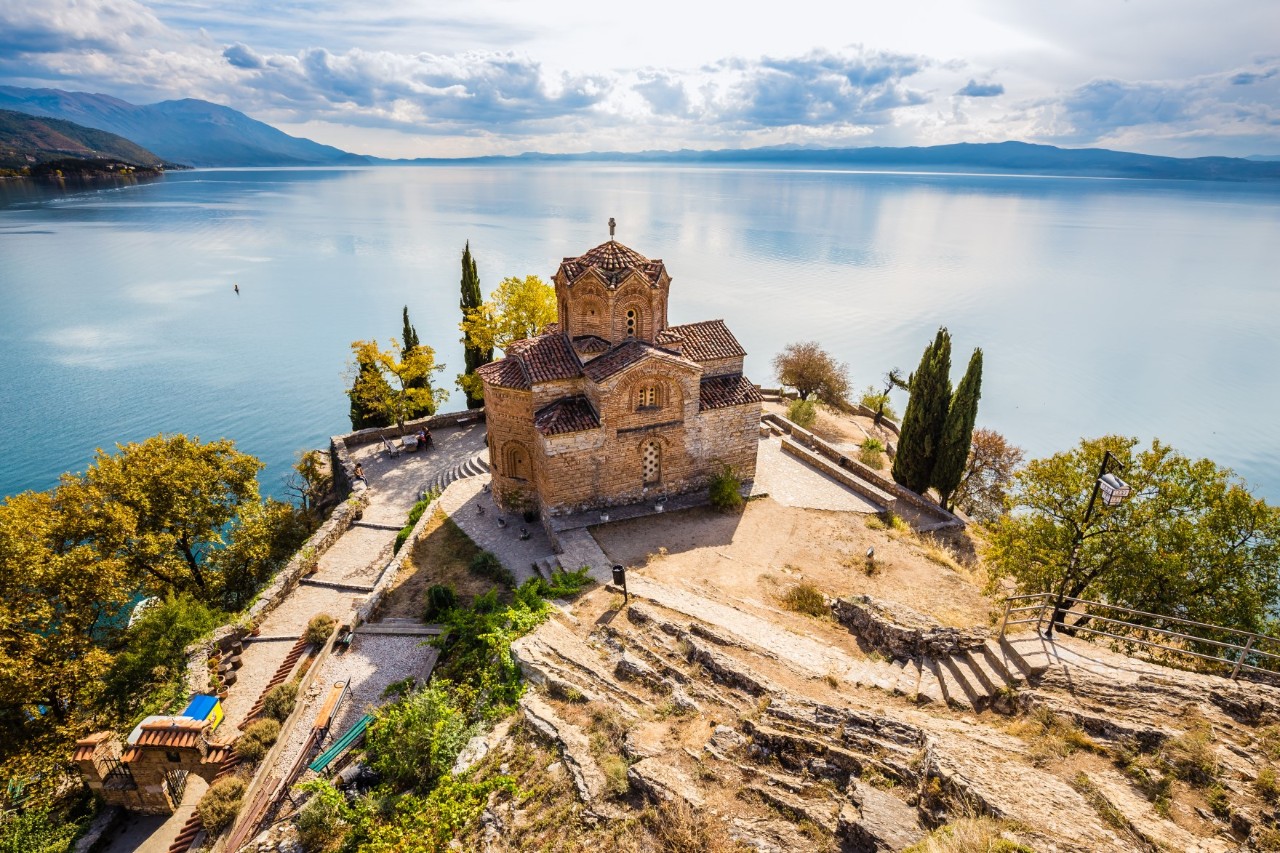 View of an Orthodox church, which stands on a cliff on a lake surrounded by flat mountains. The church is surrounded by green trees and various stone formations.