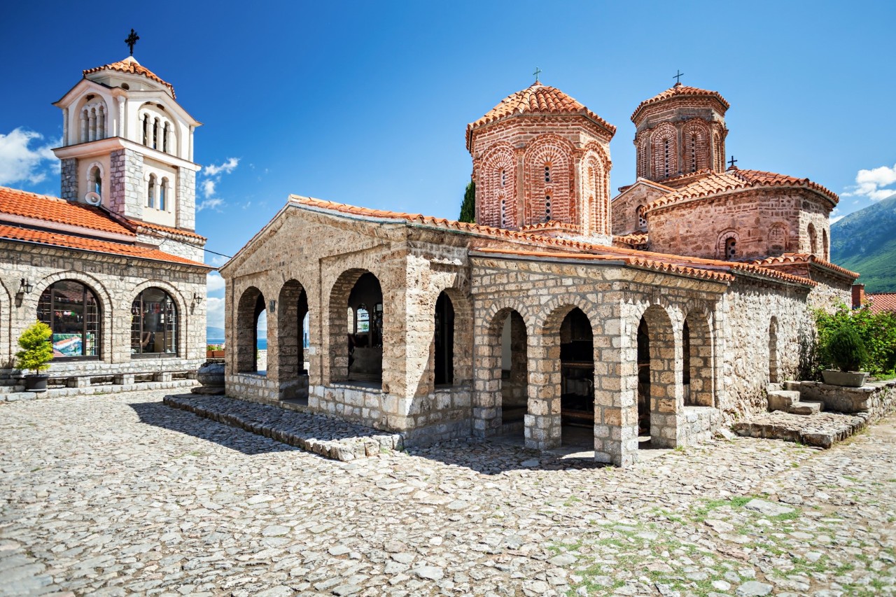 View of an impressive monastery complex surrounded by cobblestones with three towers against a blue sky. Several arched passageways can be seen on the ground floor of the monastery.