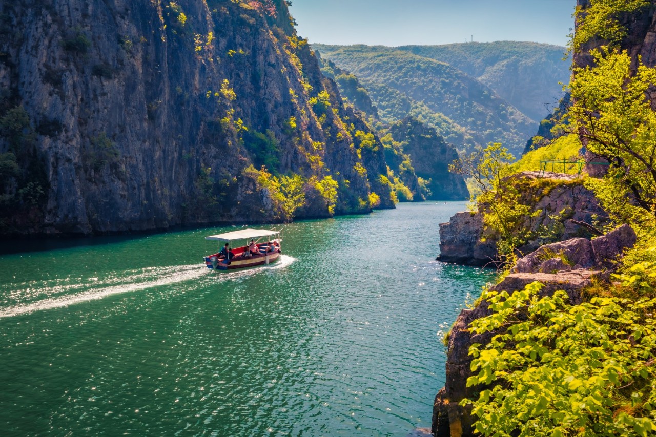 Boat with a sun sail roof on a turquoise-green river surrounded by tree-covered mountains.
