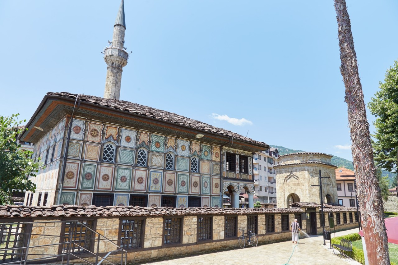 View of a mosque with various embellishments on the outer wall. There is a small wall in front of the mosque with the flat roof and a minaret can be seen behind it. Buildings and trees can be partially seen on the right.