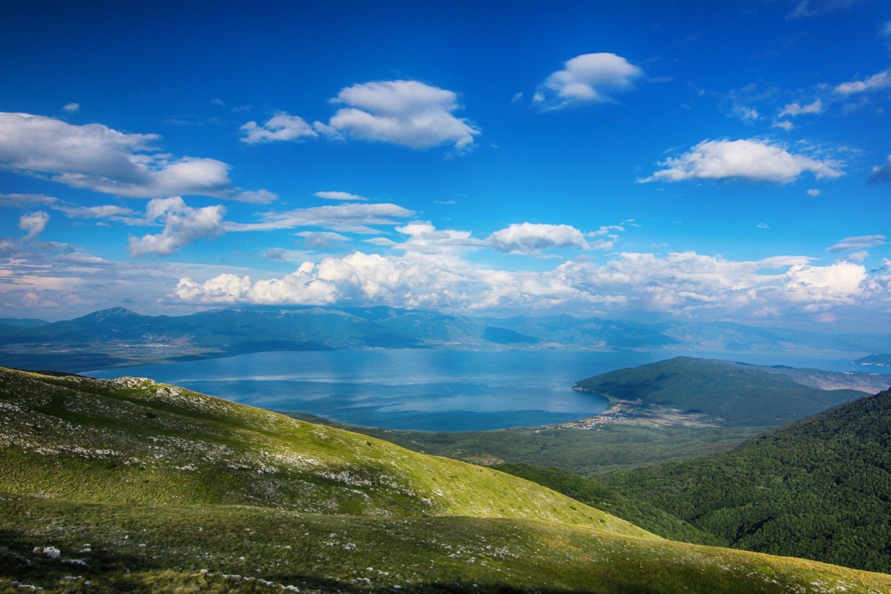 View from above of a vast, green mountain landscape in the centre of which lies a blue lake. There are clouds in the blue sky.