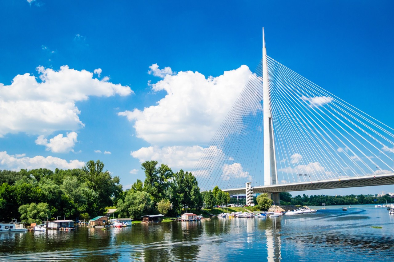 View from the water onto a modern, light-coloured steel cable bridge on the right of the picture; the steel cables converge on a high mast like a sail. On the left are trees with numerous small, colourful boats in the water in front of them.