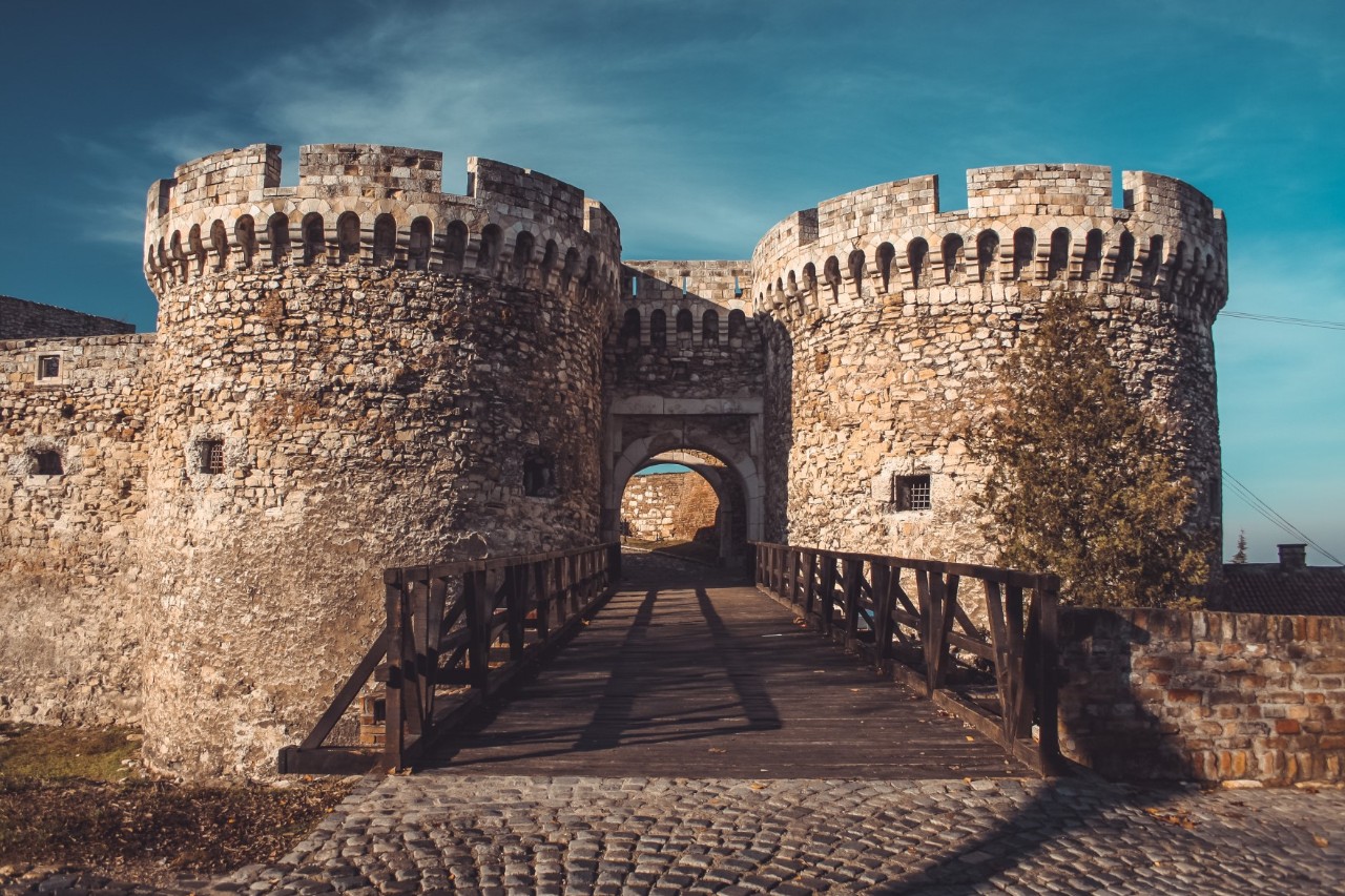 Frontal view of a castle complex with two central, very large towers, between which a wooden bridge leads through a gate into the castle. 