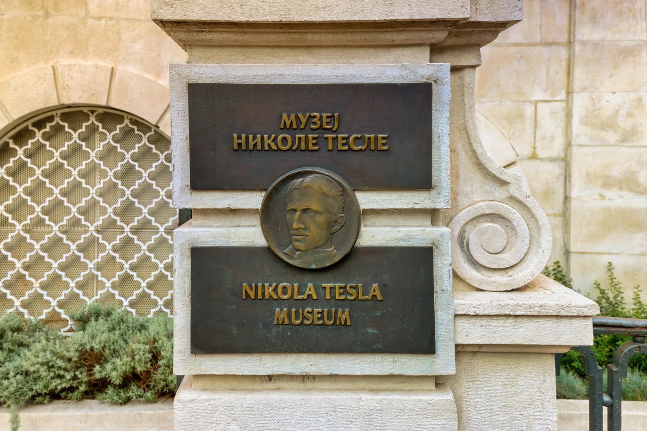 In the centre are two rectangular metal plates attached to a white, historic building. The panels are connected by the portrait of a man, also set in metal. The lower plate reads “Nikola Tesla Museum”, and the upper plate is written in Cyrillic.