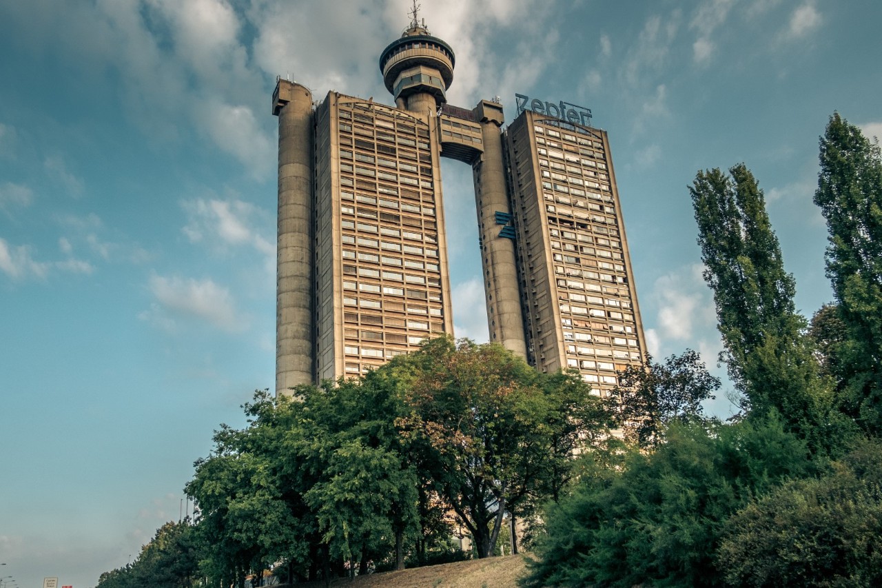 A two-part high-rise building, which is connected by two column-like elements and a spire. There is a large gap between a pillar and one tower block. There are trees in front of the building.