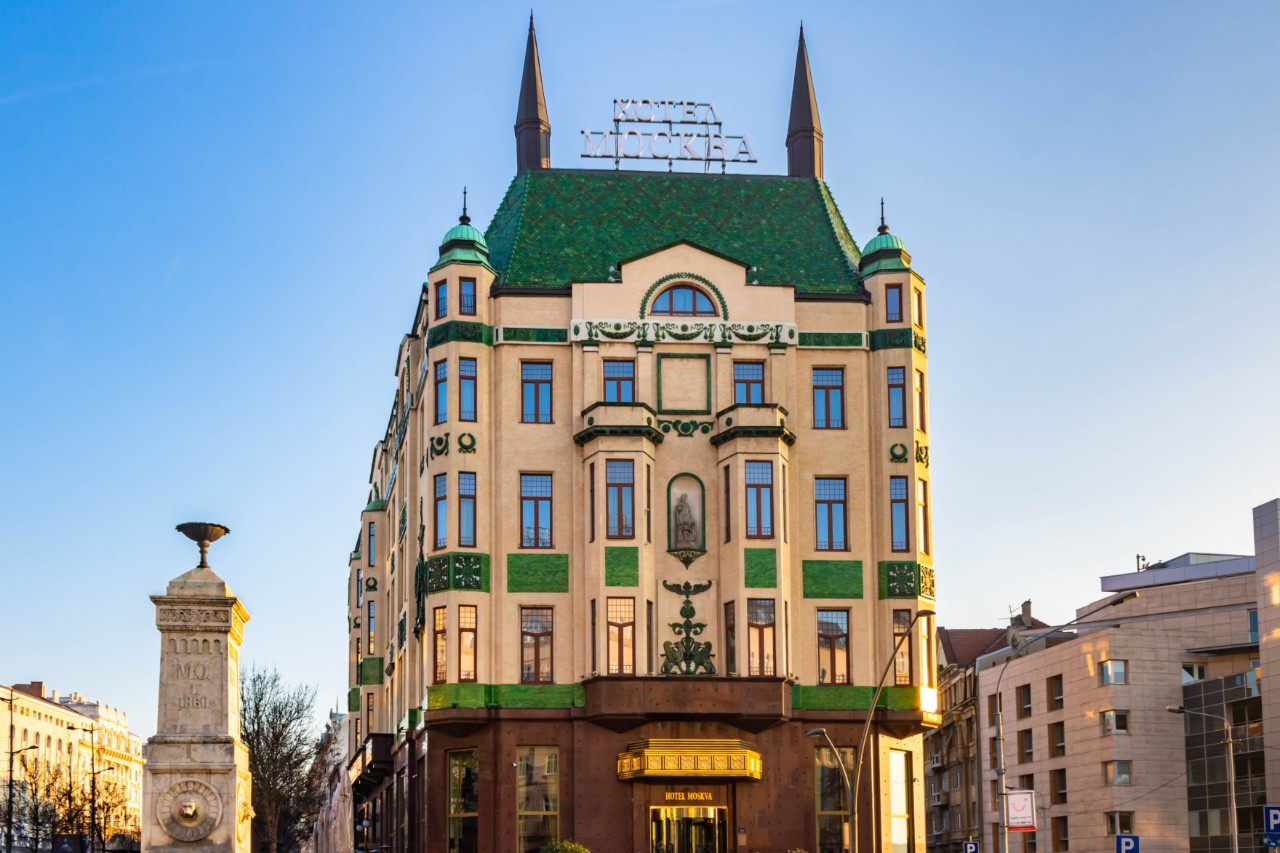 View of a five-storey art nouveau building decorated with turrets and bay windows. The front is sand-coloured with green elements and a green roof on which two more towers stand. To the left of the building is a smaller, white tower, and to the right a modern building.