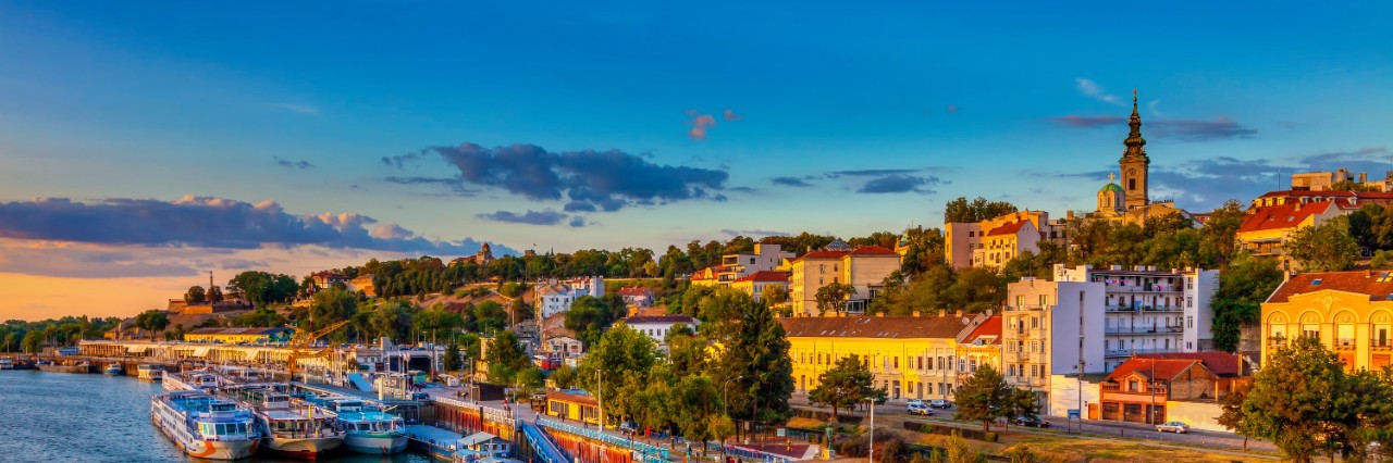 View from above of a harbour on a riverbank in the evening light. Several excursion boats are moored in the harbour. The riverbank is lined with trees and there are buildings behind it and a church tower in the distance.