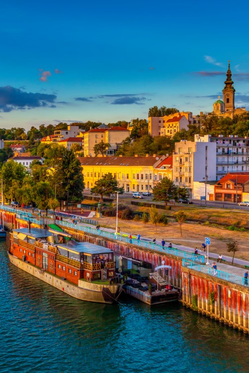 View from above of a harbour on a riverbank in the evening light. An excursion boat is moored in the harbour. The riverbank is lined with trees and there are buildings behind it and a church tower in the distance.