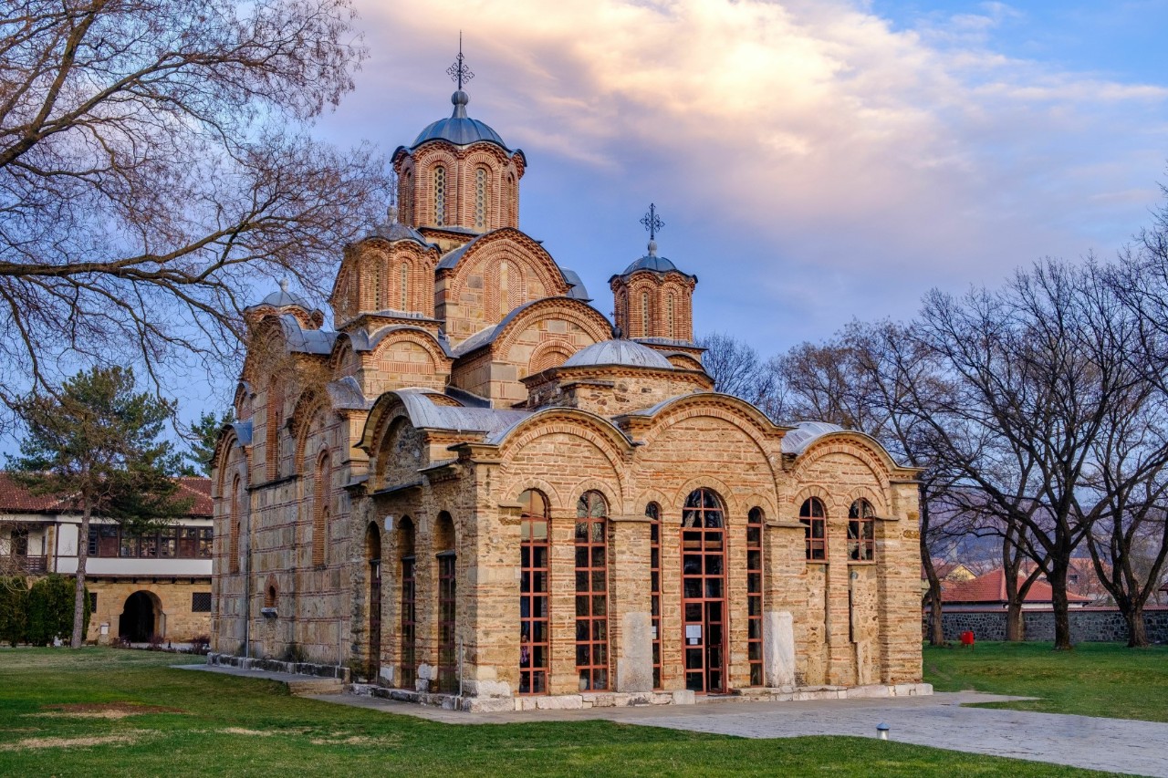 A large brick church with tall, arched windows that glow red in the evening light. The church is made up of numerous rounds, domed building elements. A cross is mounted on each of the two highest domed towers. The building is surrounded by a lawn and a small paved square, with a few leafless trees standing around it.