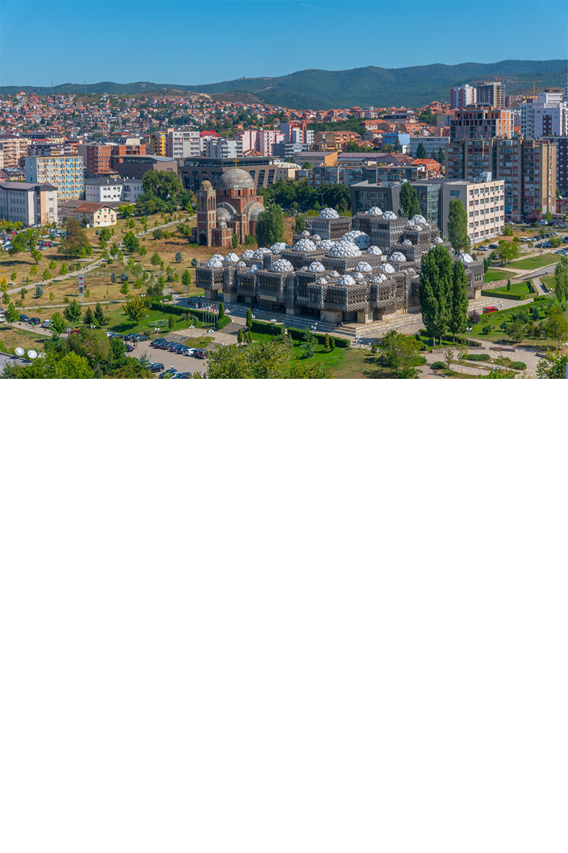 View of the city from above. In the foreground, green lawns, trees, and several paved areas can be seen. Closest to the viewer is a large, jagged, grey structure with numerous dome-shaped towers, behind which rise many multi-storey houses. A chain of hills can be seen in the background.