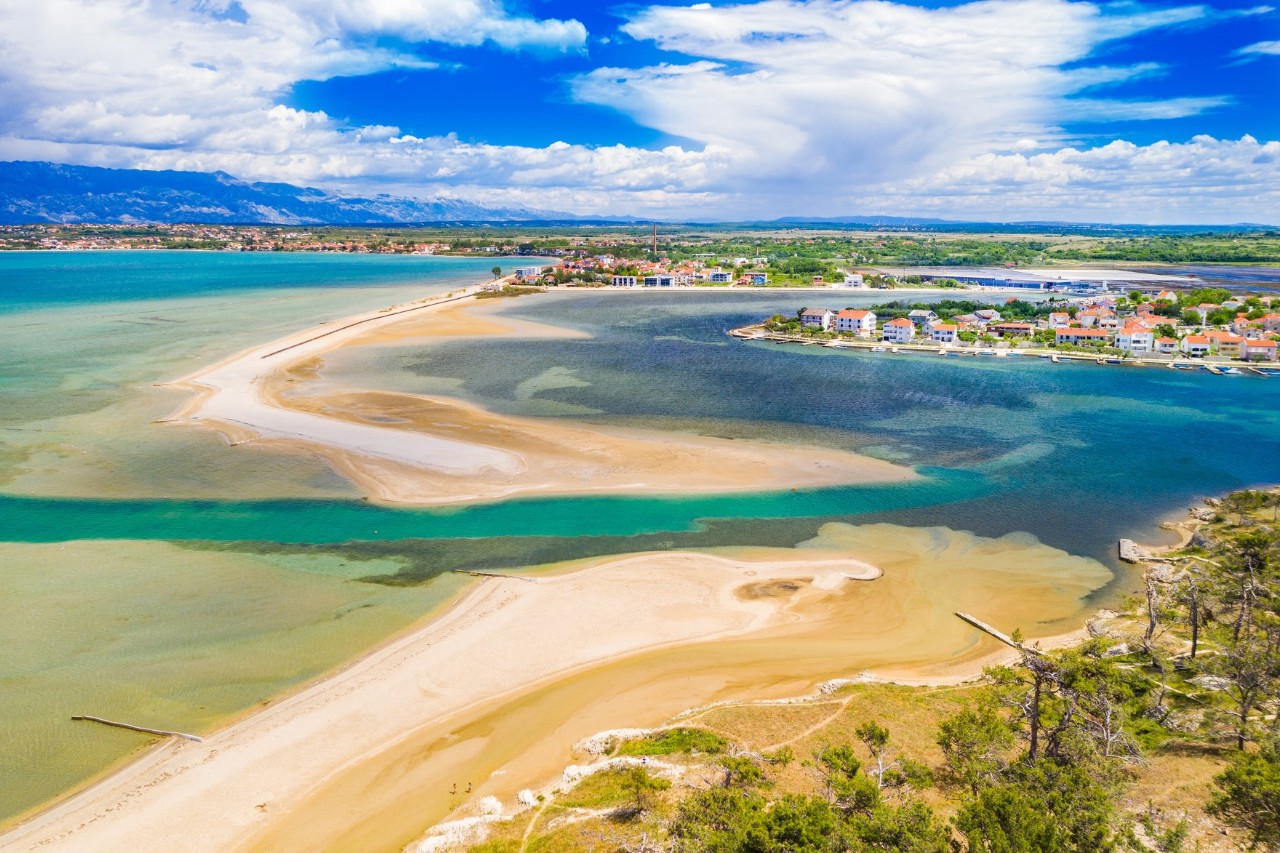 Aerial view of a lagoon beach. A large, light-coloured sandbank separates the sea on the left from the lagoon on the right. The lagoon and the sea are connected by a turquoise-coloured arm of water. A few trees can be seen in the foreground. A small island with houses lies in the lagoon.