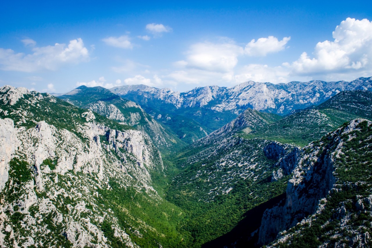 Panoramic view of a mountain range with various rock formations, some of which are covered in green. Several clouds are visible in the blue sky above the peaks.