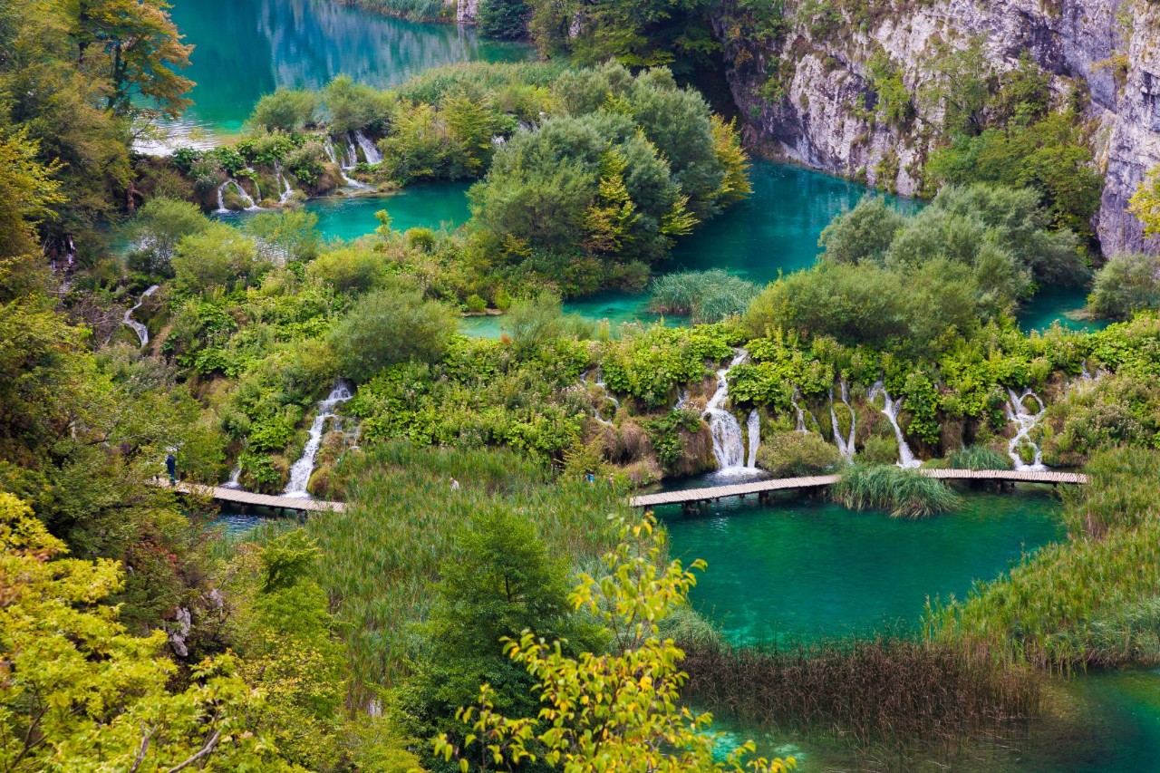 Aerial view of several turquoise-blue lakes connected by waterfalls. A dense forest can be seen between the lakes and the waterfalls. A steep rock face rises on the right-hand side of the picture.
