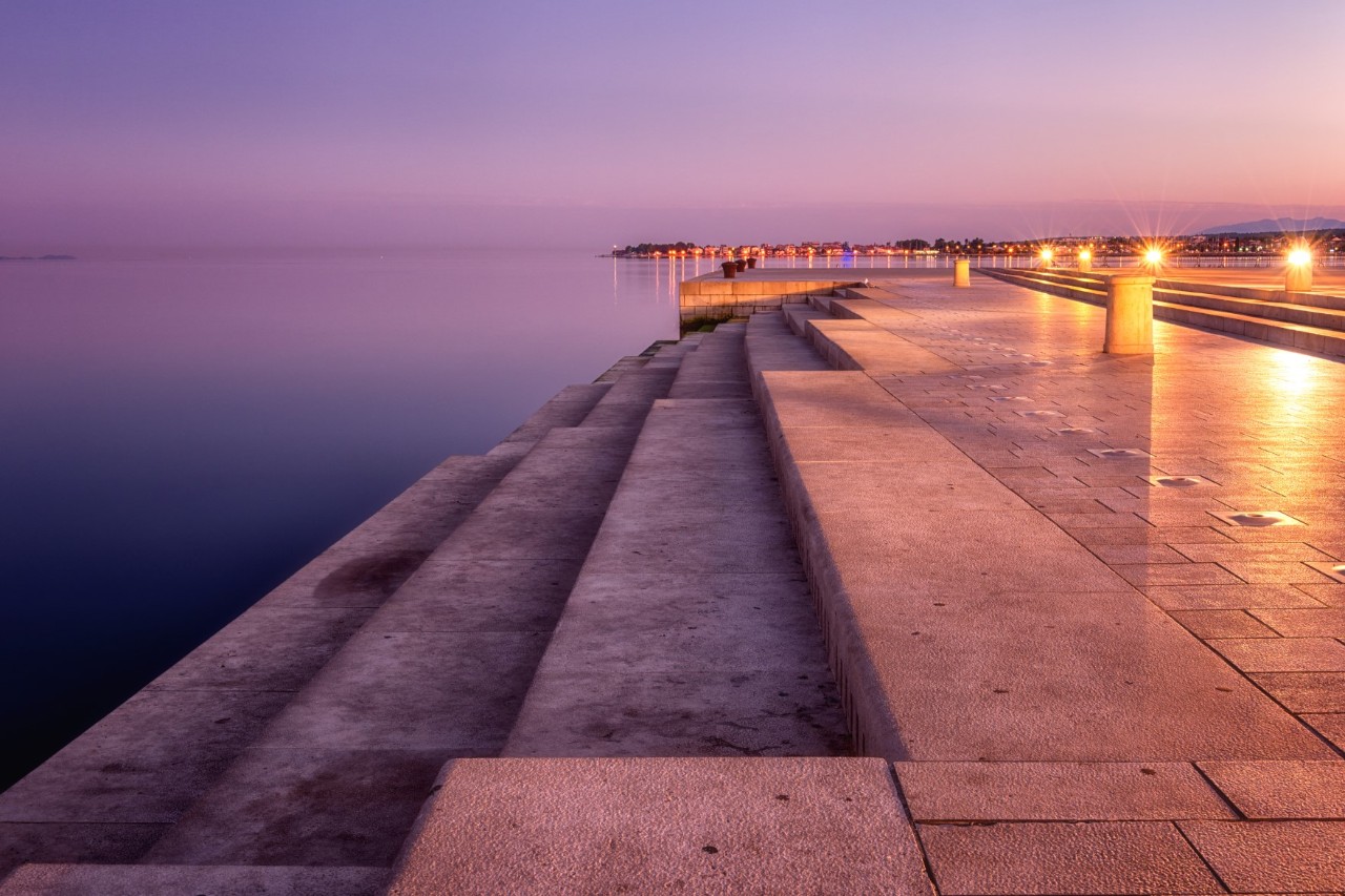 A wide stone staircase leads directly to the sea, the water and steps glowing in the purple-coloured evening sky. The illuminated city can be seen far in the background.
