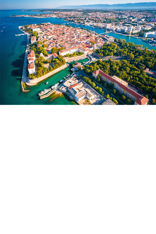 Aerial view of a coastal town. In the centre of the picture is a densely built-up, almost rectangular peninsula surrounded by turquoise blue water. The houses have red tiled roofs. The peninsula is connected to the mainland, which is densely forested. In front of the forest in the foreground is a very long building with a red, triangular tiled roof. In the background you can see the rest of the town, which is connected to the peninsula by a bridge.