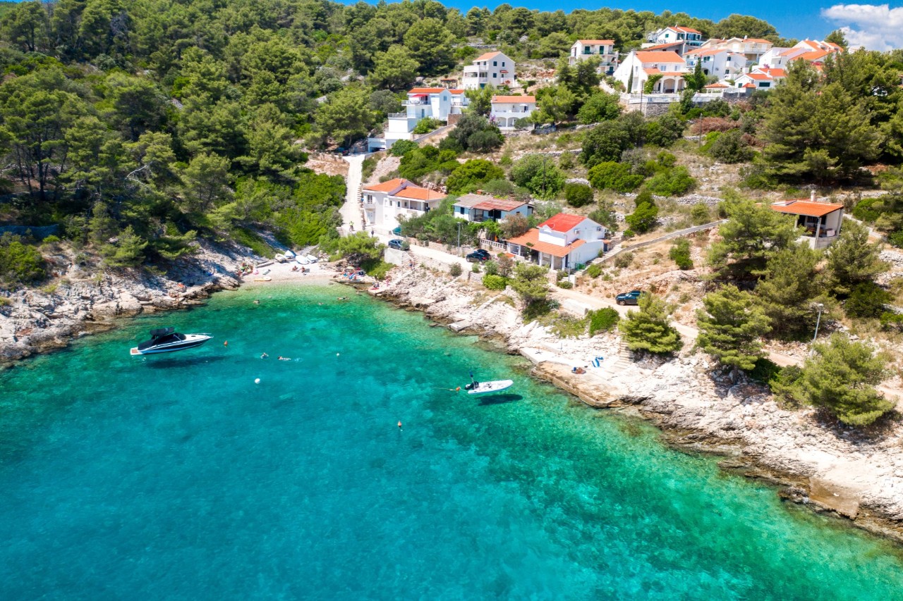 The picture shows a small bay with crystal clear water. Along the rocky coastline are picturesque houses with red roofs, surrounded by greenery and pine trees. Two small boats are anchored in the water. 