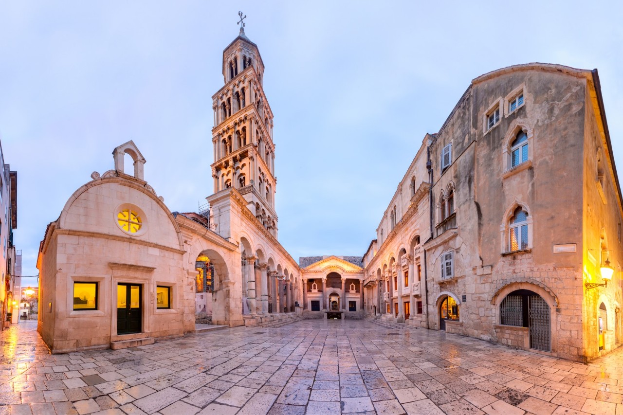 A historic building complex made of light-coloured stone with a striking bell tower in the twilight. The ensemble is surrounded by a clean, smooth stone floor.