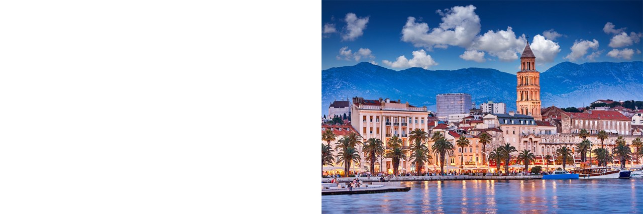 old town at dusk. In the foreground, there is a built-up waterfront promenade with boats, palm trees, cafés and people. A bell tower rises above it. Behind it, there is a mountain range. 