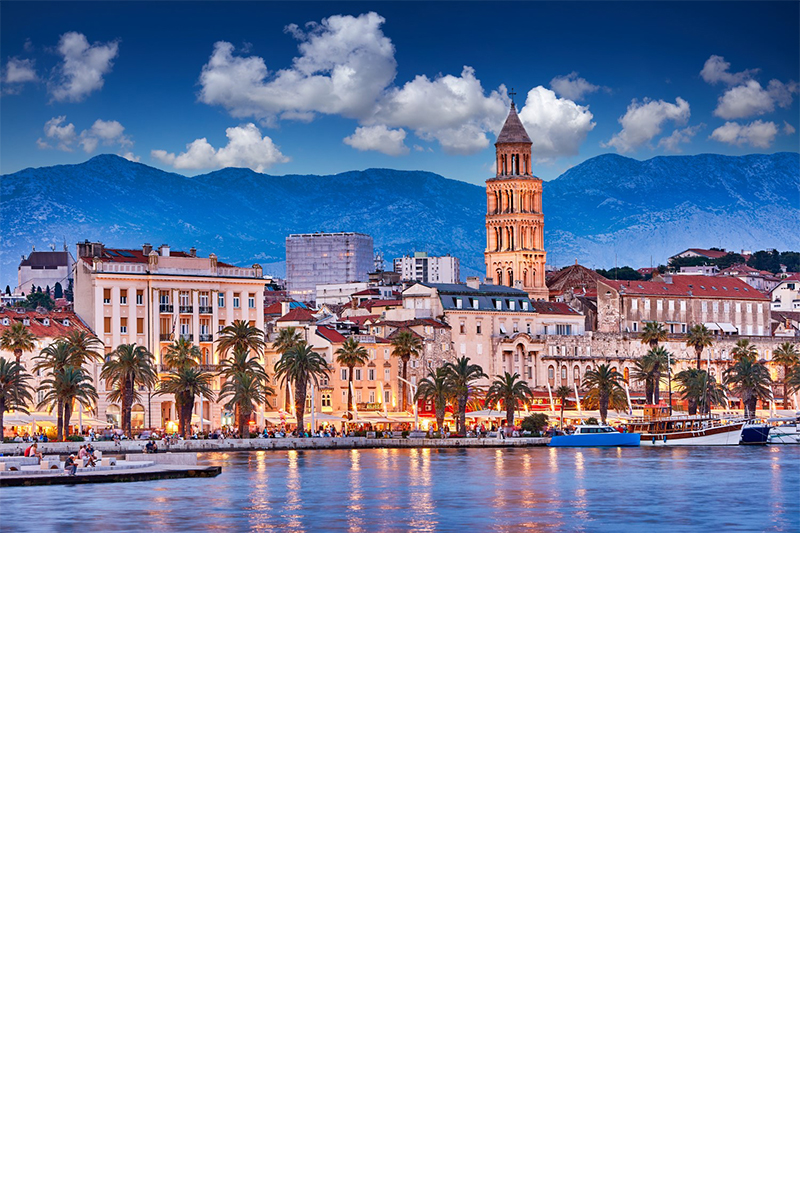 old town at dusk. In the foreground, there is a built-up waterfront promenade with boats, palm trees, cafés and people. A bell tower rises above it. Behind it, there is a mountain range. 