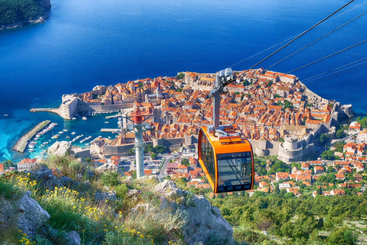 Bird’s-eye view of an old town jutting out into the sea, surrounded by a city wall. In the foreground is an orange-coloured cable car gondola. It looks as though the picture was taken from high up from a cable car. There are more buildings outside the city wall. Densely overgrown rocks can be seen in the foreground.