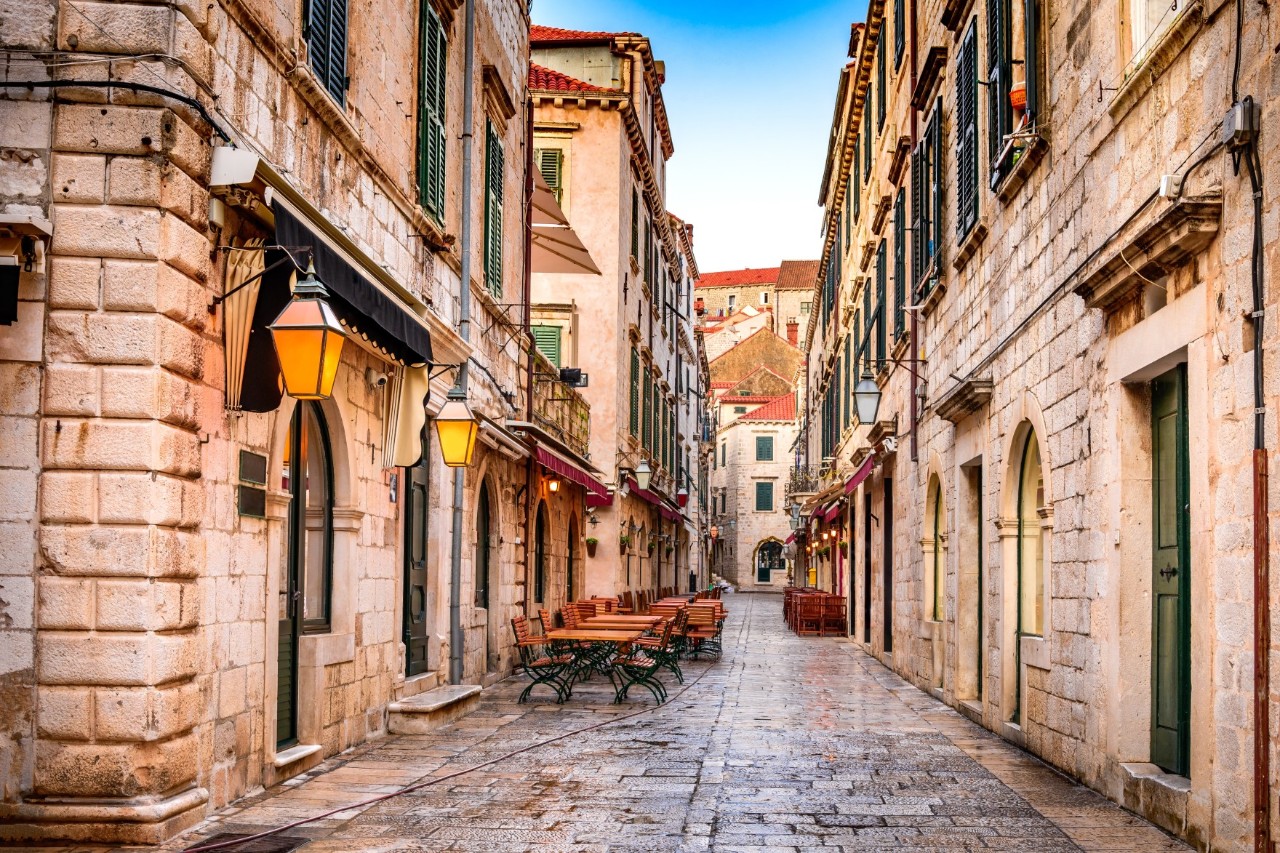 View into an old town street; there are empty tables and chairs in front of several restaurants. Three-storey old buildings with green window shutters rise on either side. Illuminated historical lanterns hang from the building on the left of the picture. The roads are wet, the sky is blue. It has been raining.