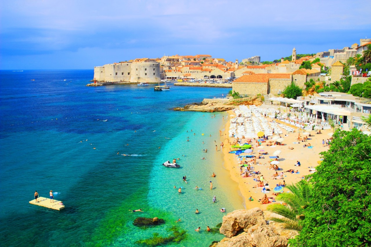 A small, yellow sandy beach with white sunbeds, parasols and lots of people. The turquoise sea is crystal clear. Some people are swimming in the water. On the left is a larger raft with two people. Old town buildings with red-tiled roofs and a fortress can be seen in the background.