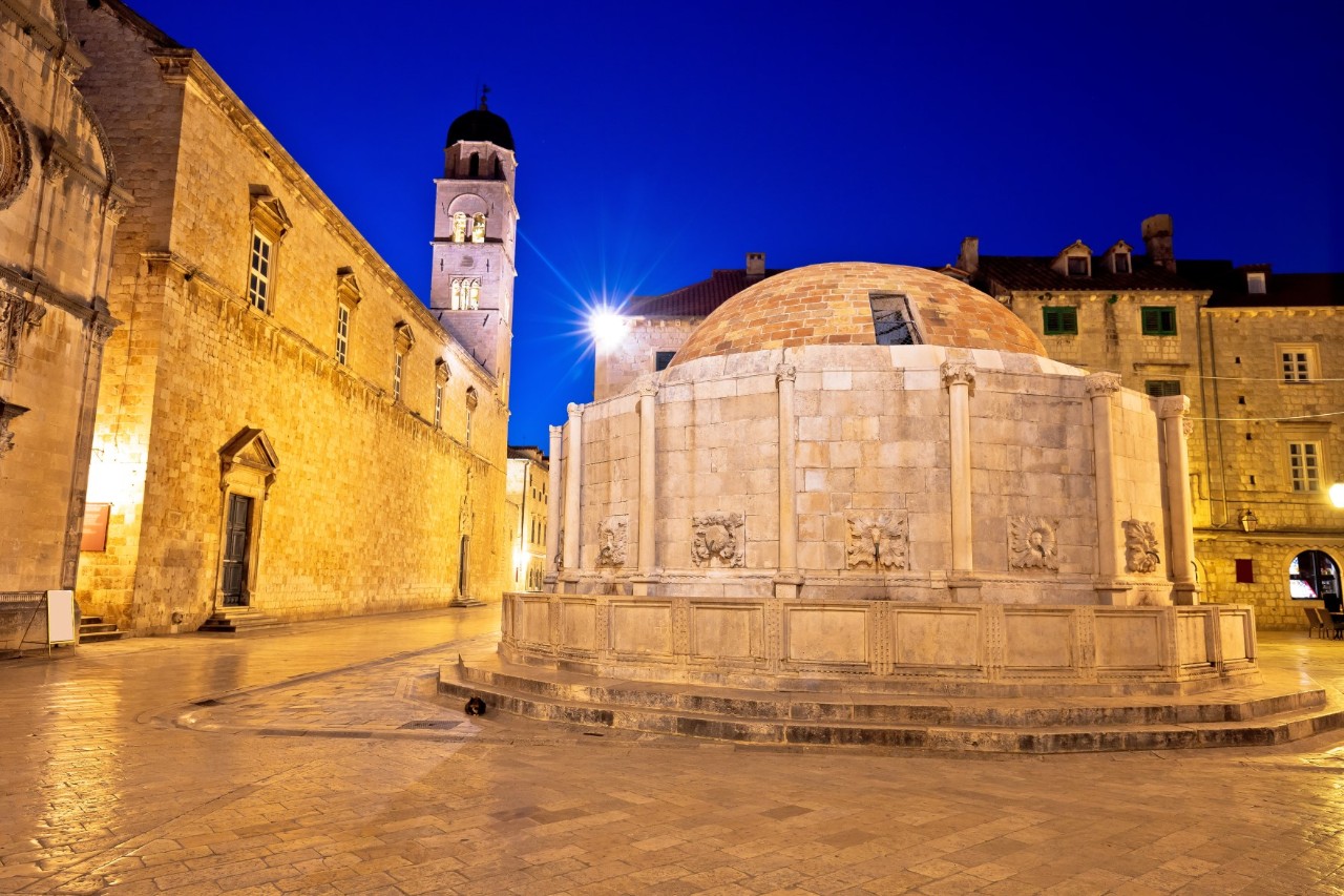 A picture taken in the evening of a large, round fountain on a brightly paved square in an old town. The fountain is surrounded by a high wall and covered by a dome. Three steps lead to the basin in front of the fountain. To the left of the fountain is the long nave and narrow tower of a church, which is illuminated.